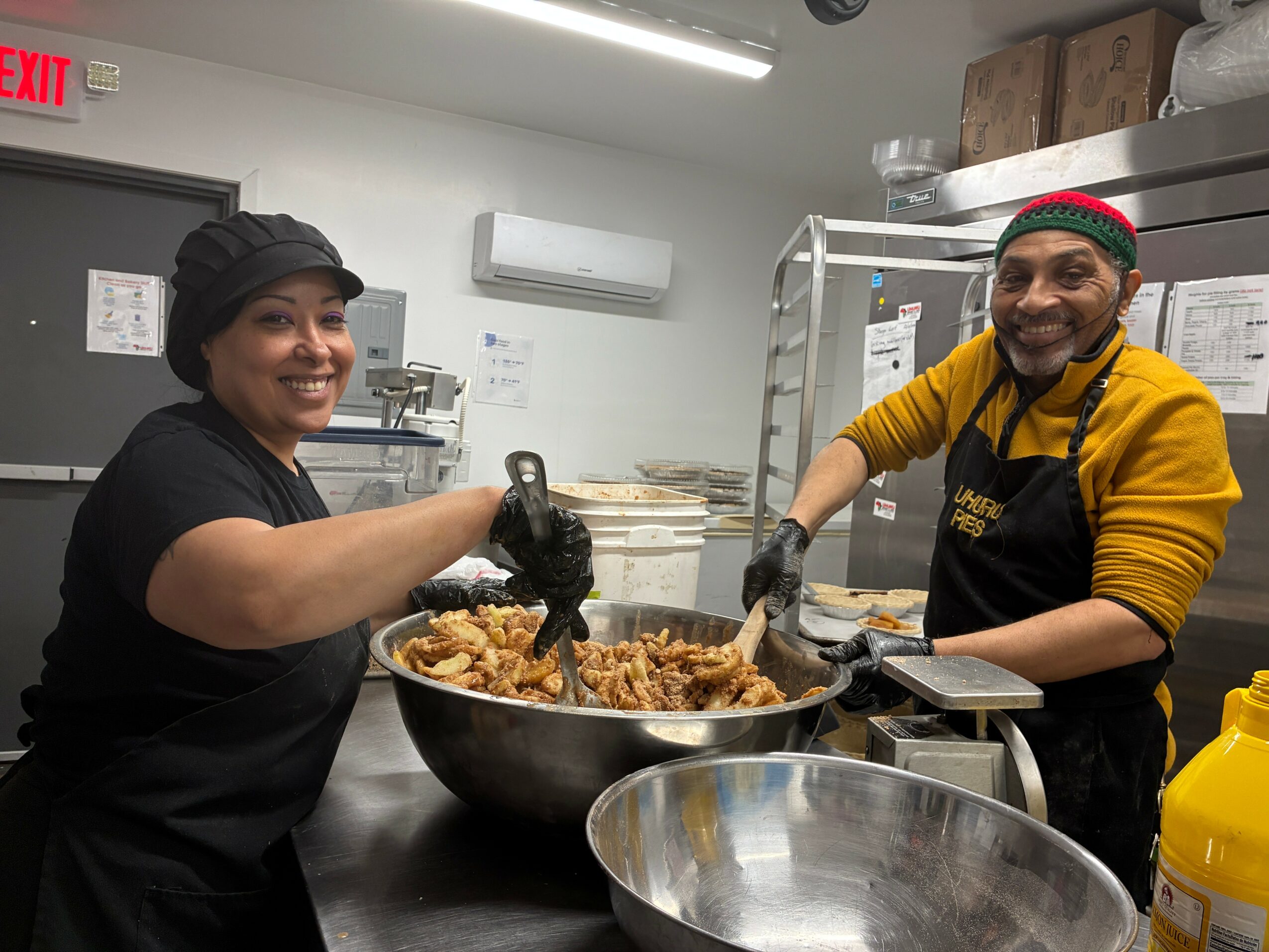 Two people cooking together, stirring a large pan of food in a kitchen, smiling at the camera.