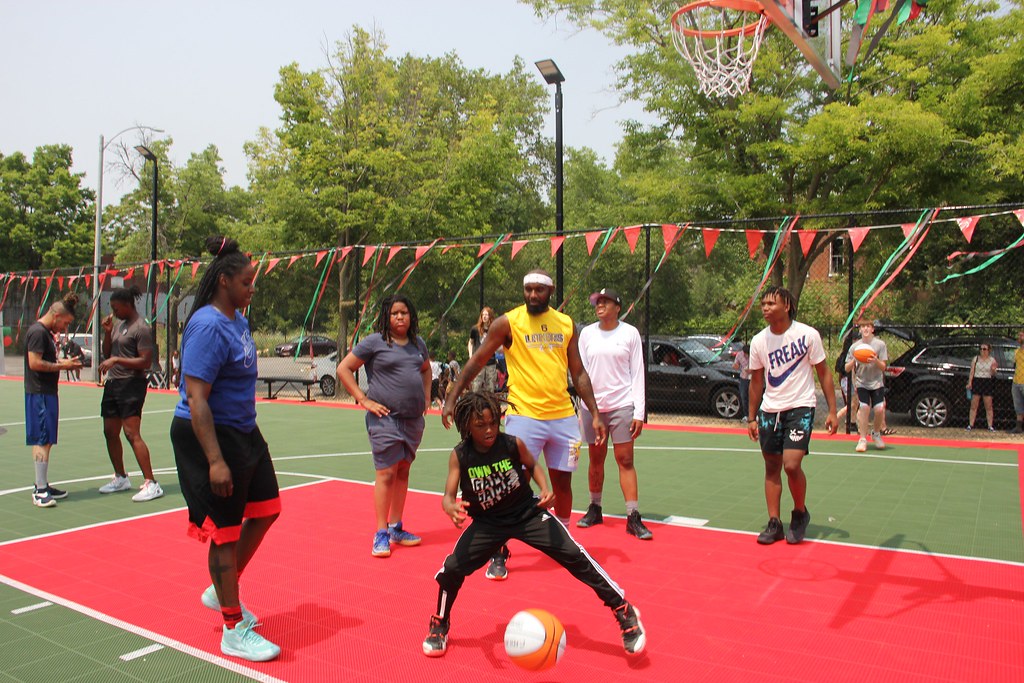 Children playing basketball on an outdoor court with trees and parked cars in the background.