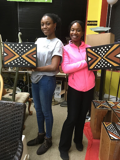 Two women standing indoors, each with a laptop, smiling, with colorful walls and furniture in the background.