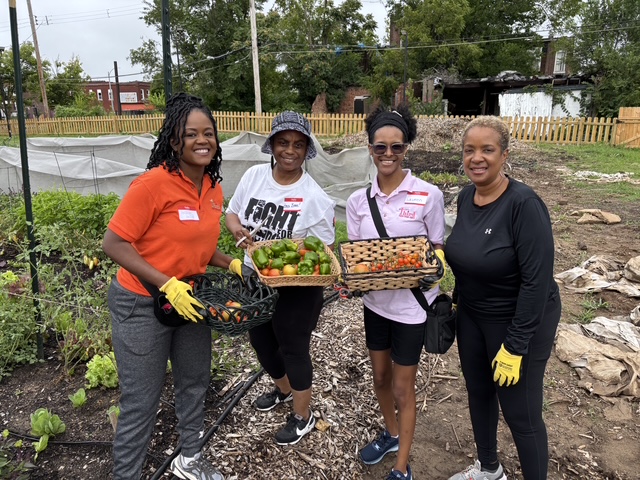 Four women standing in a garden holding baskets of vegetables, smiling, with trees and a fence in the background.