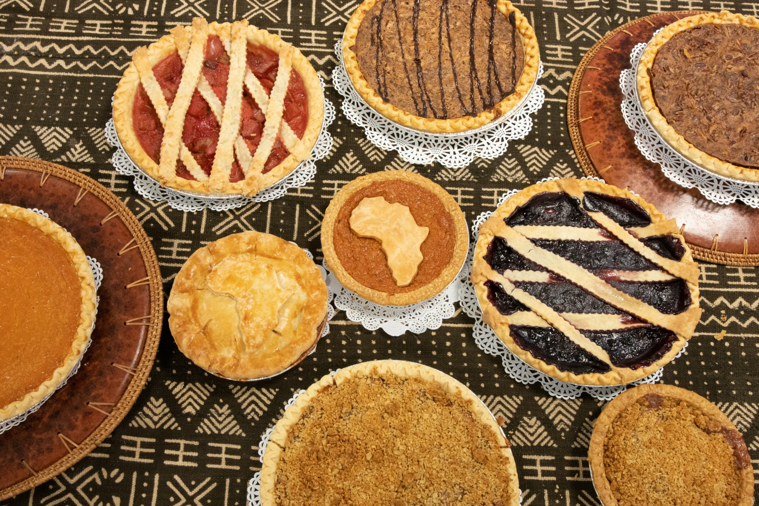 Assorted cookies and pastries on a patterned surface, including a lattice-topped tart, round cookies, and a pastry with chocolate drizzle.