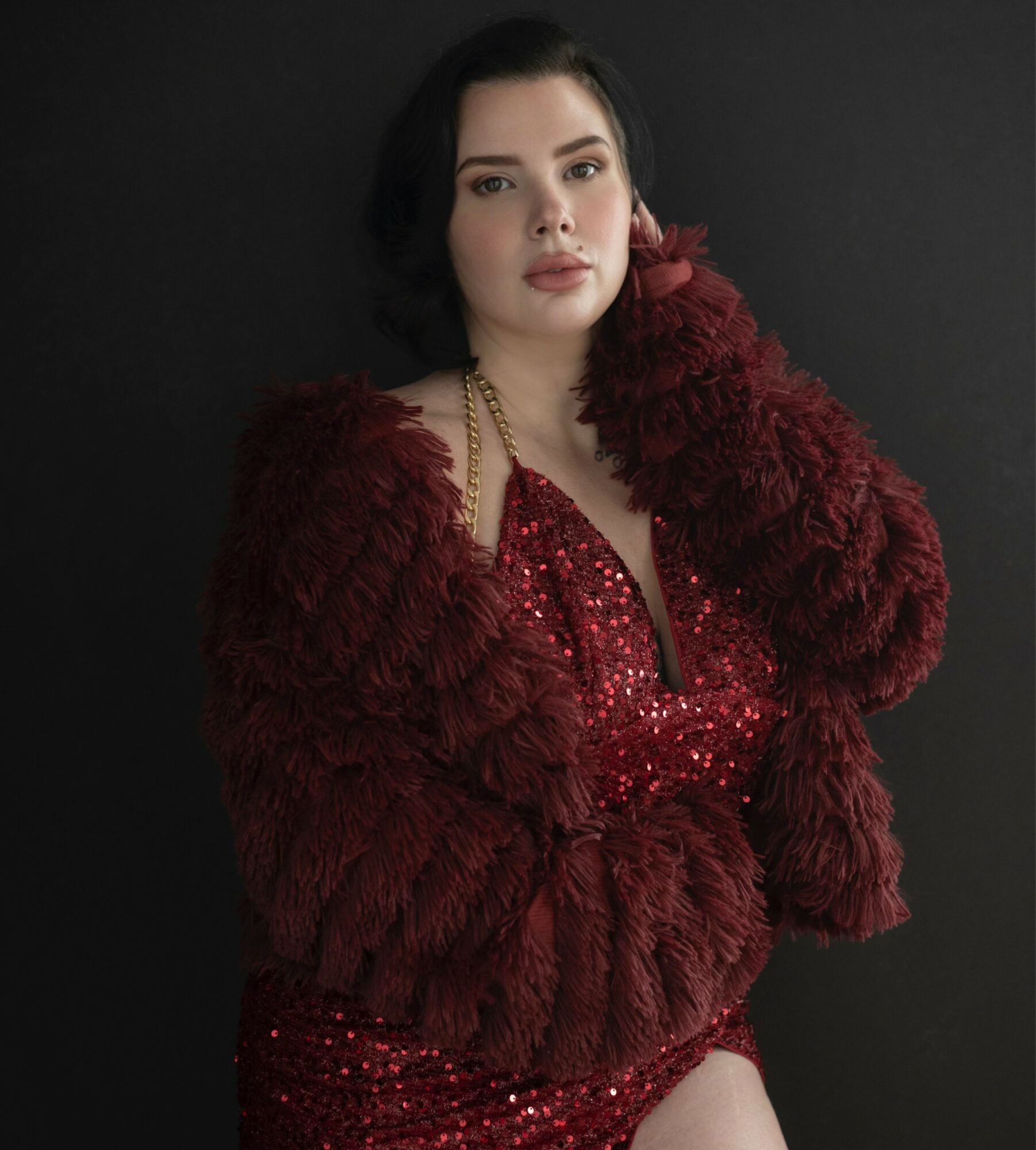 Woman with dark hair in a red sequined dress and a red fur coat against a black background.
