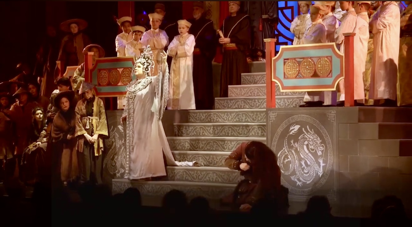 Stage scene with people in historical costumes, some on stairs, one kneeling, ornate decorations, and a dark background.