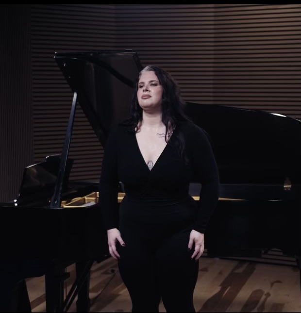 Woman in black standing in front of a grand piano in a music studio, with wooden wall panels behind her.