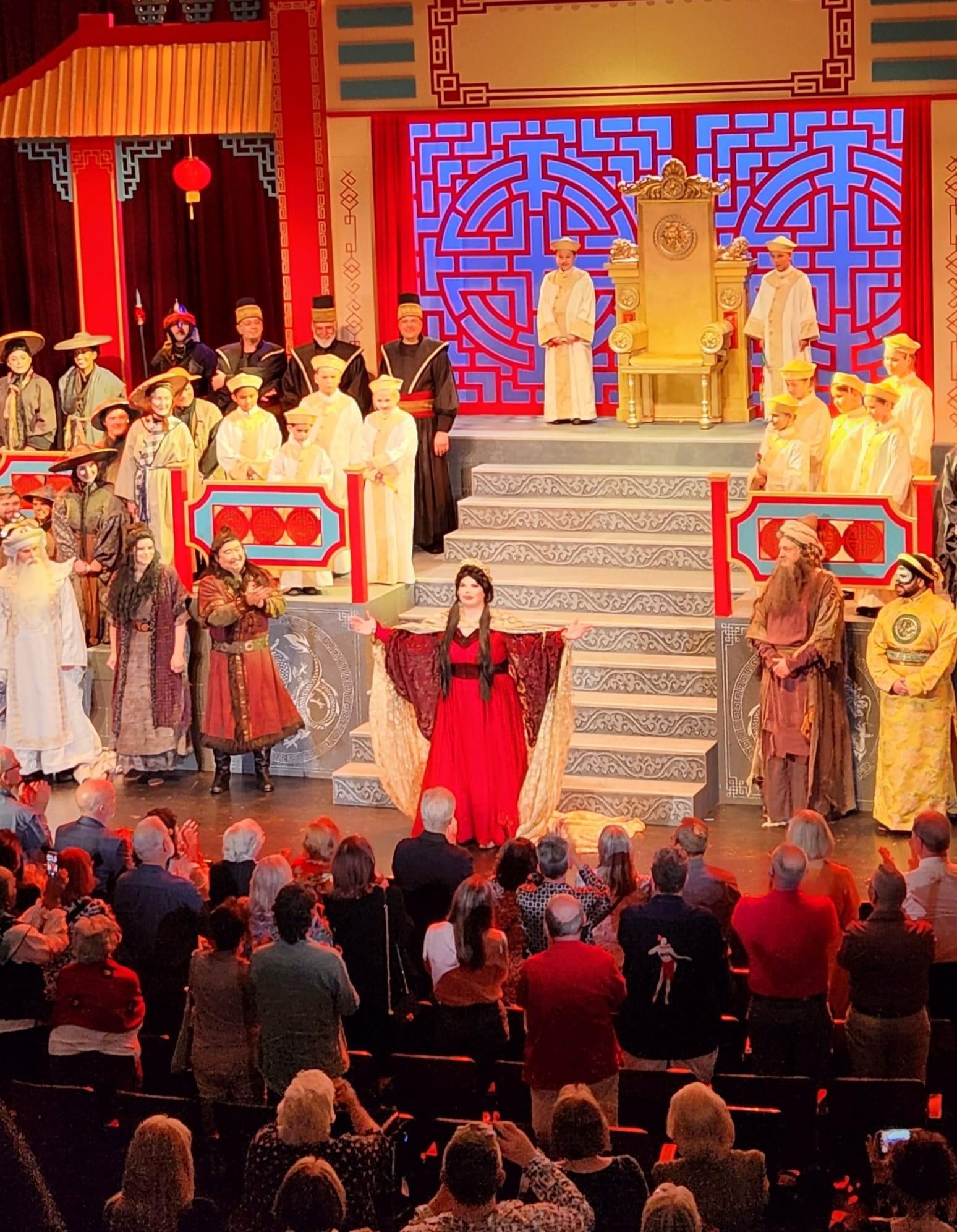 The stage features performers in traditional costumes, with a woman in a red dress at the center, and an audience watching.