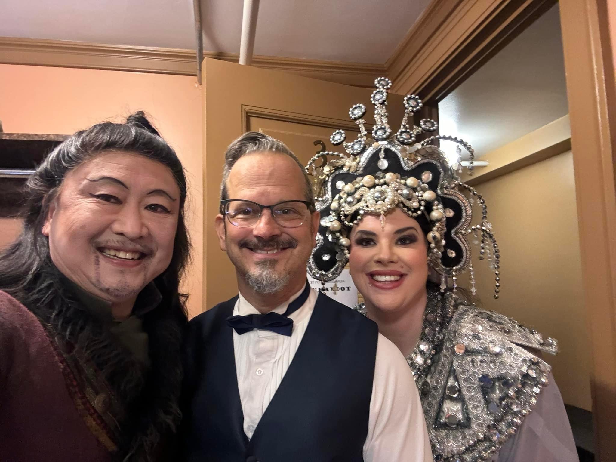 Three people smiling indoors, one in traditional costume with elaborate headpiece, two men in formal attire.