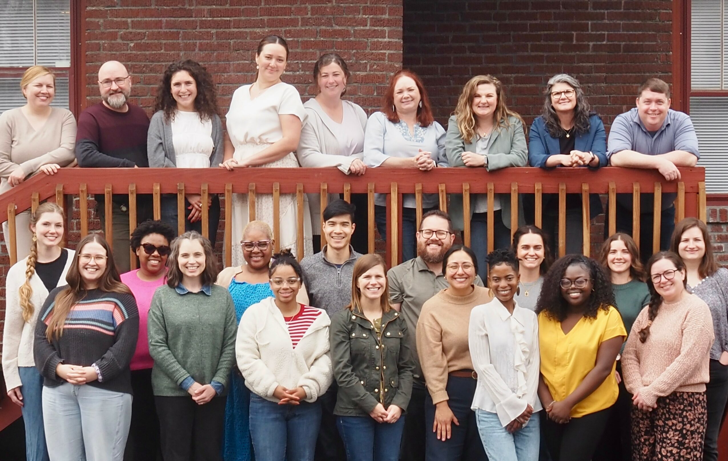 Group of 20 people standing outdoors on a wooden balcony, smiling, with a brick building in the background.