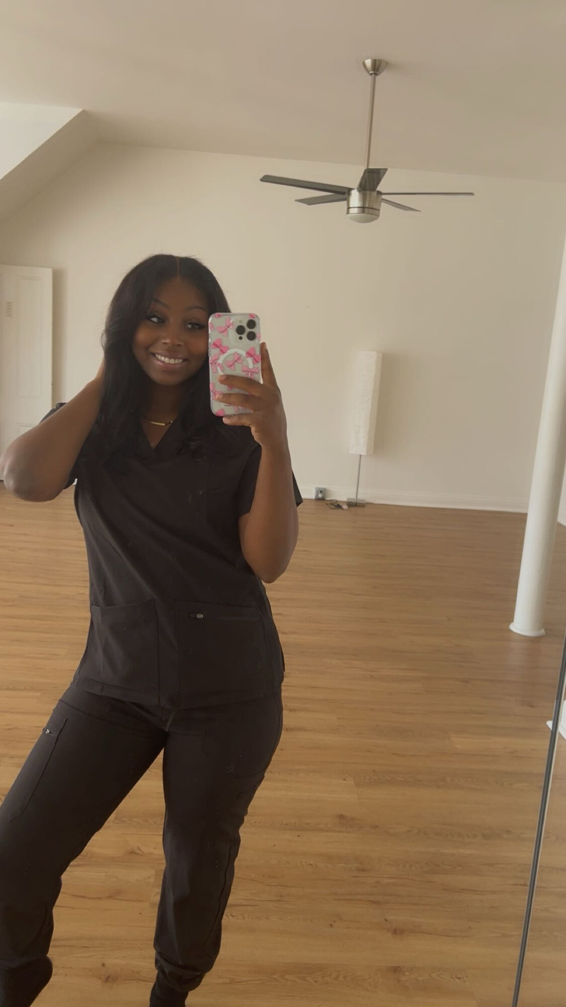 Woman taking a mirror selfie in a room with wooden floor, ceiling fan, and white walls.