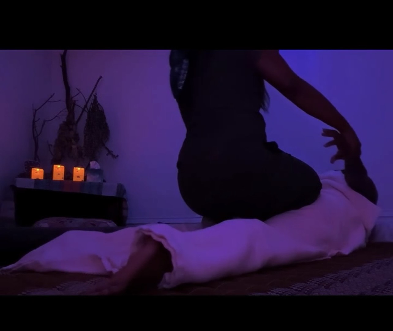Person kneeling on a person lying on the floor in a dimly lit room with candles and decorative branches in the background.