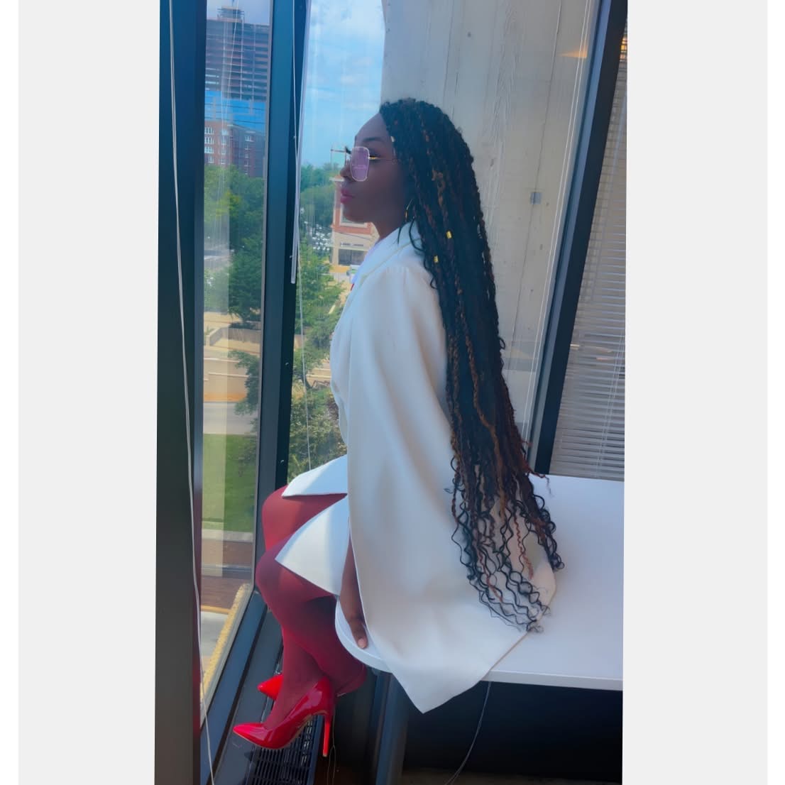 Person with long braided hair sitting by a window, looking outside, wearing a white top, with cityscape view.