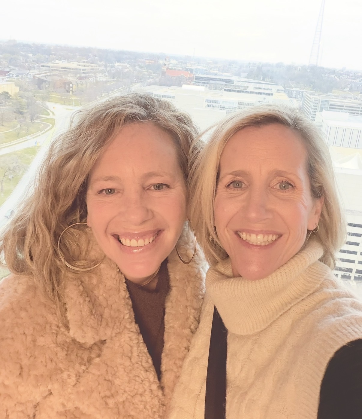 Two women smiling outdoors with cityscape background, one wearing a beige coat, the other in a cream turtleneck.