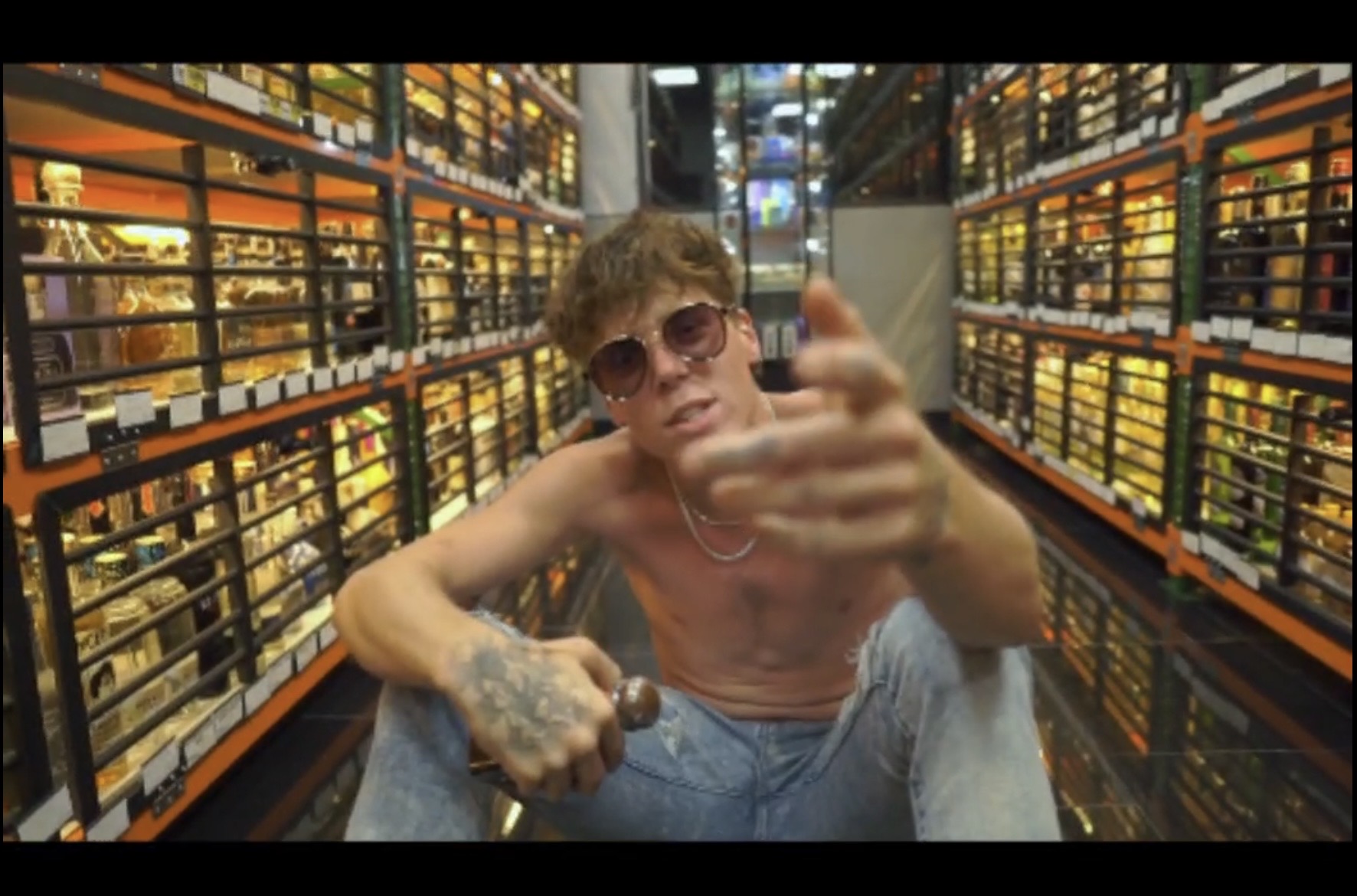 Young man with sunglasses, shirtless, pointing at the camera in a store aisle with shelves of products.