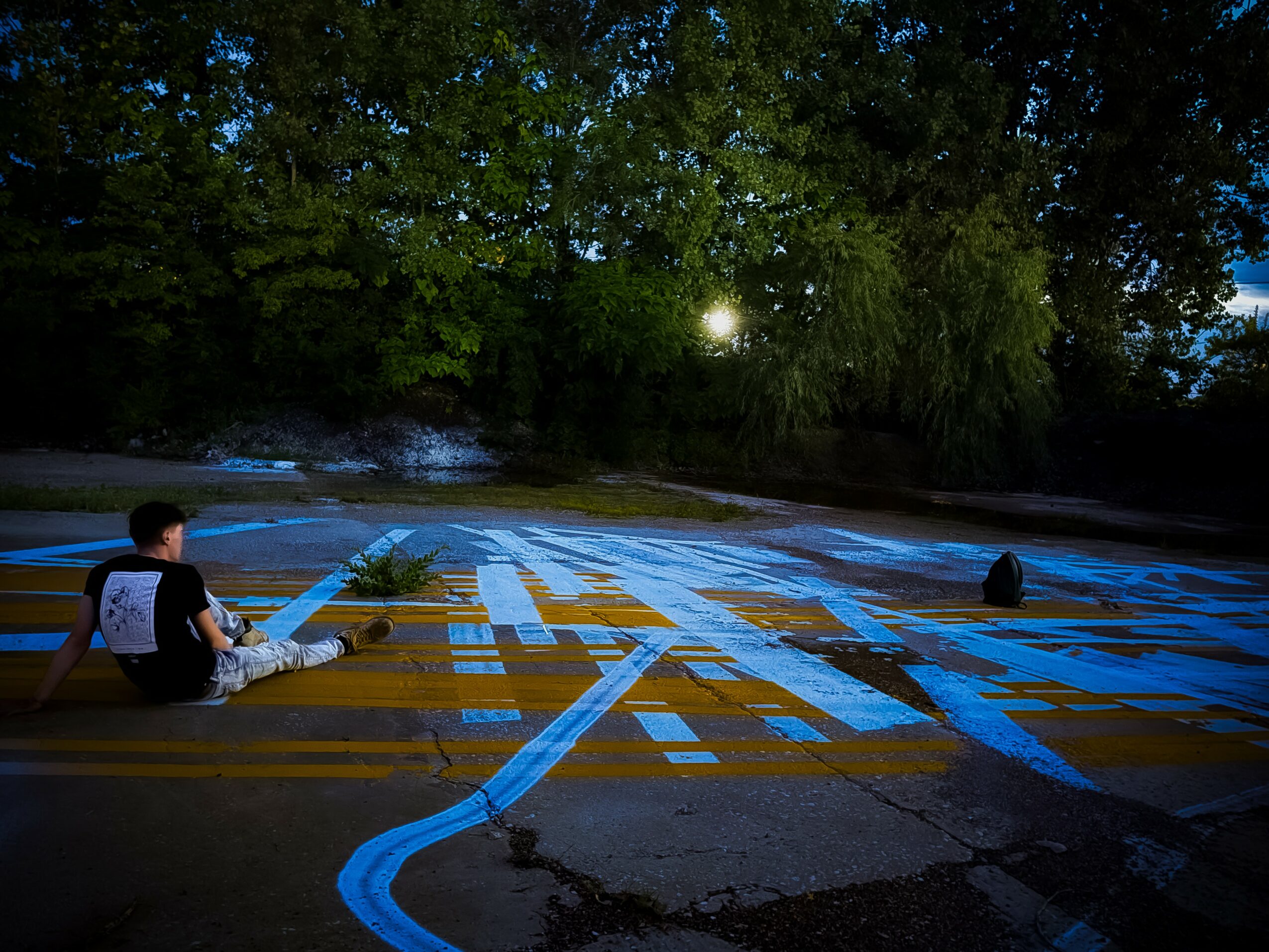 Two children sit on a crosswalk at dusk, with trees and the moon in the background, and painted lines on the road.