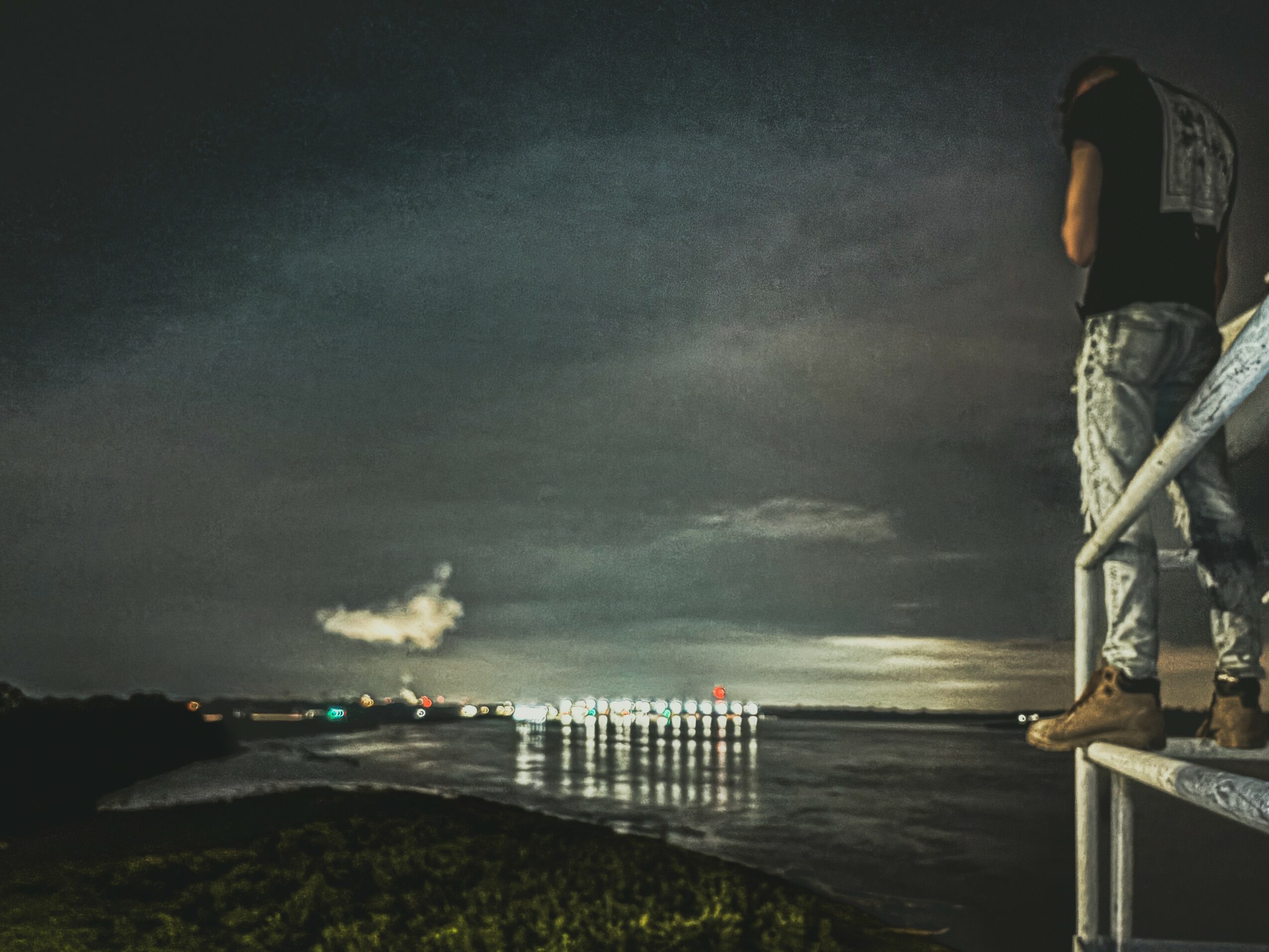 Person standing on a railing overlooking a body of water at night with city lights in the distance.