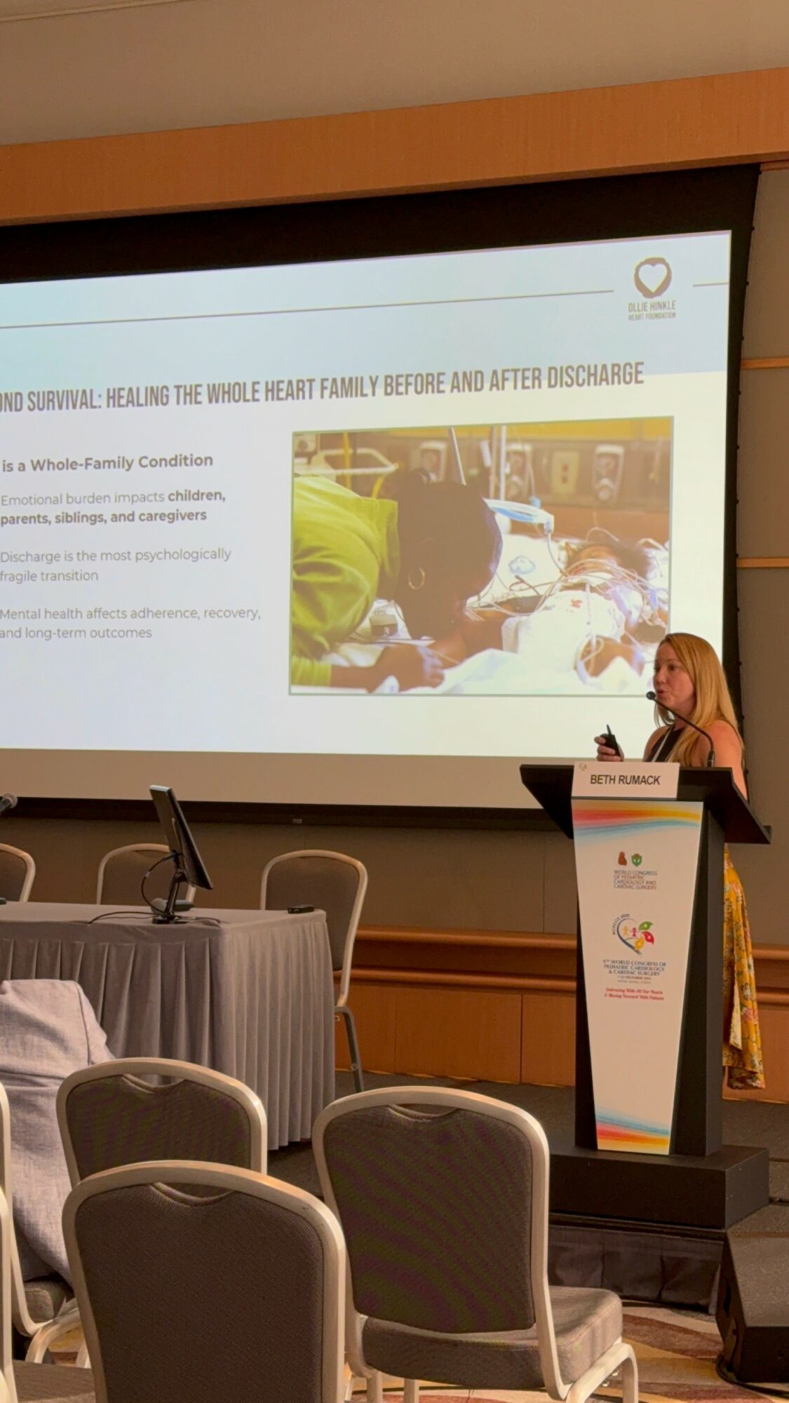 A woman stands at a podium giving a presentation in a conference room with chairs and a large screen displaying a slide with text and an image.