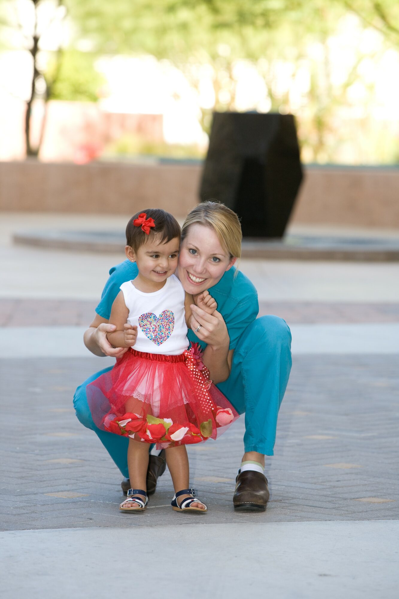 Smiling woman crouching next to a young girl outdoors, both looking at the camera, with trees and a black structure in the background.
