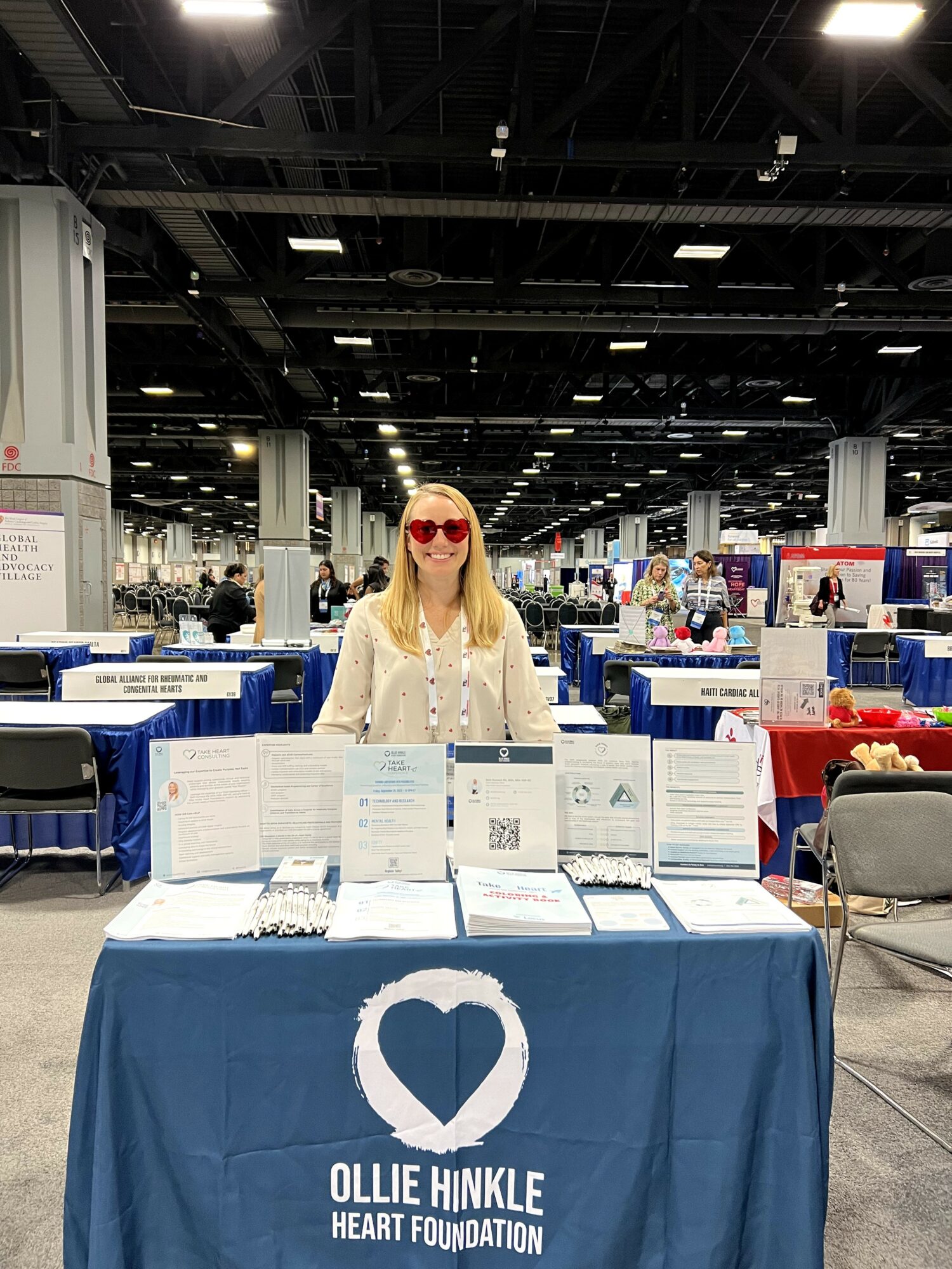 Woman with blonde hair and red sunglasses standing behind a table with informational materials at an indoor event.