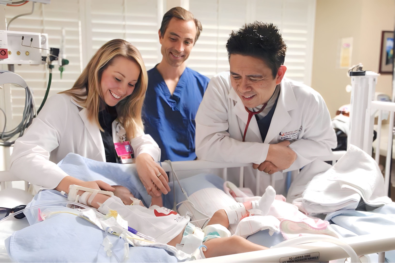 Three medical professionals smiling and interacting with a patient in a hospital room, with medical equipment visible.