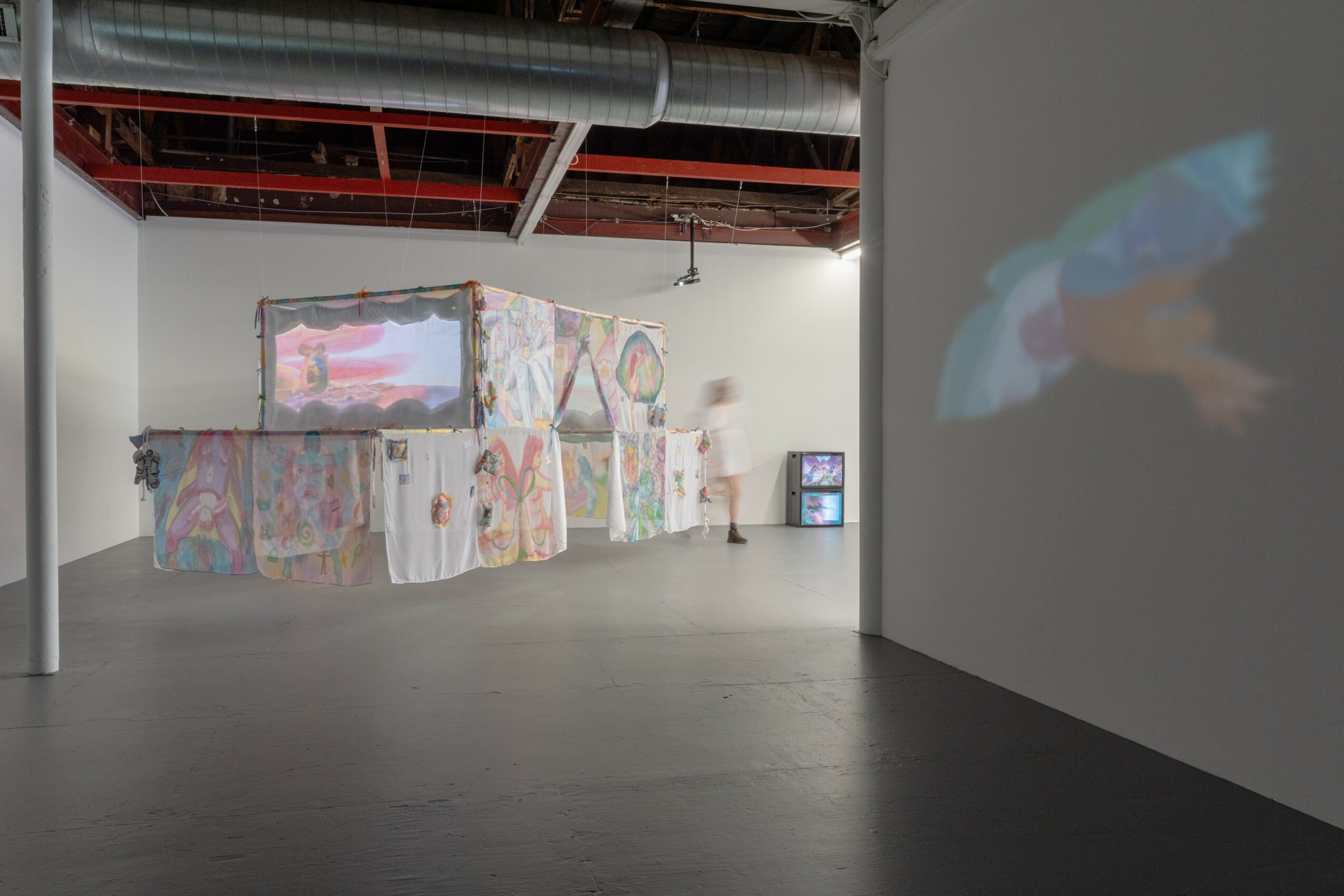 Gallery space with colorful abstract art installation on wall, two screens displaying visuals, and a person observing art.