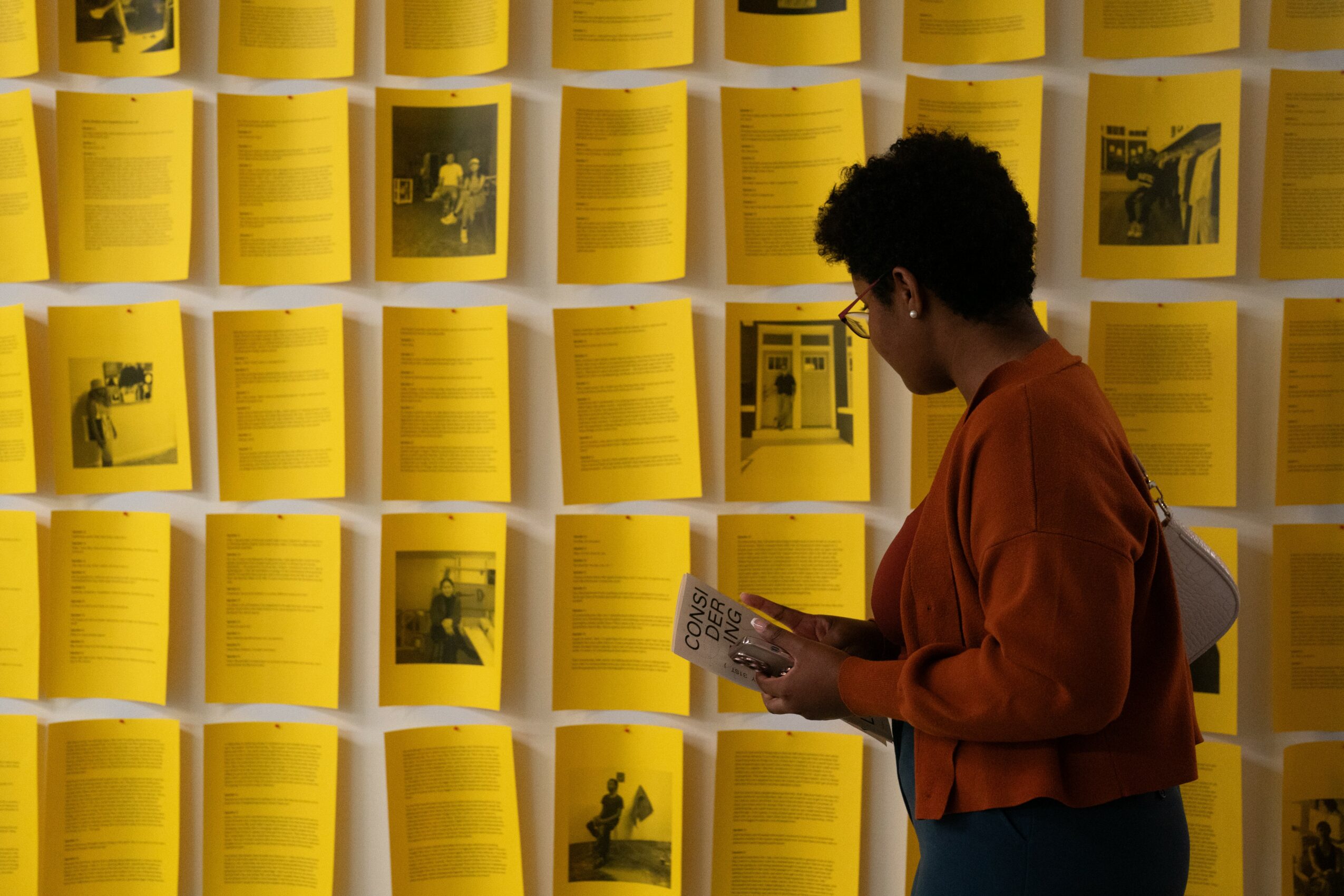 Person with short curly hair and glasses reads a booklet in front of a wall covered with yellow papers and photos.