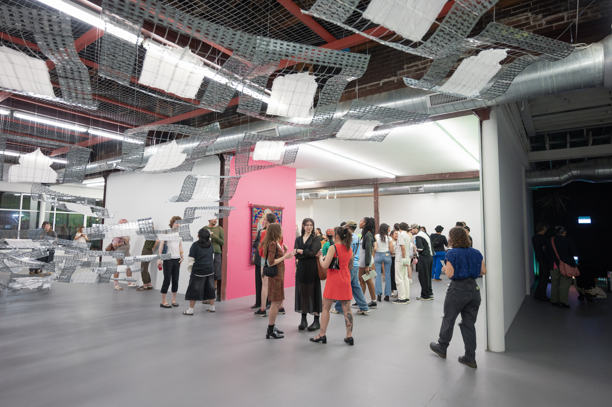 People gather in a modern indoor space with industrial ceiling elements and a pink wall, some waiting in line.