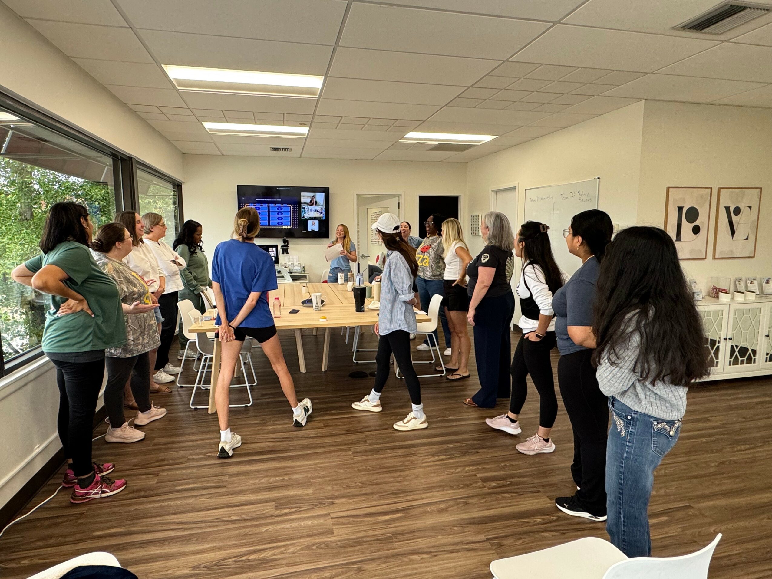 Group of people standing in a room with wooden floor, white walls, and a large screen on the wall, engaging in an activity.