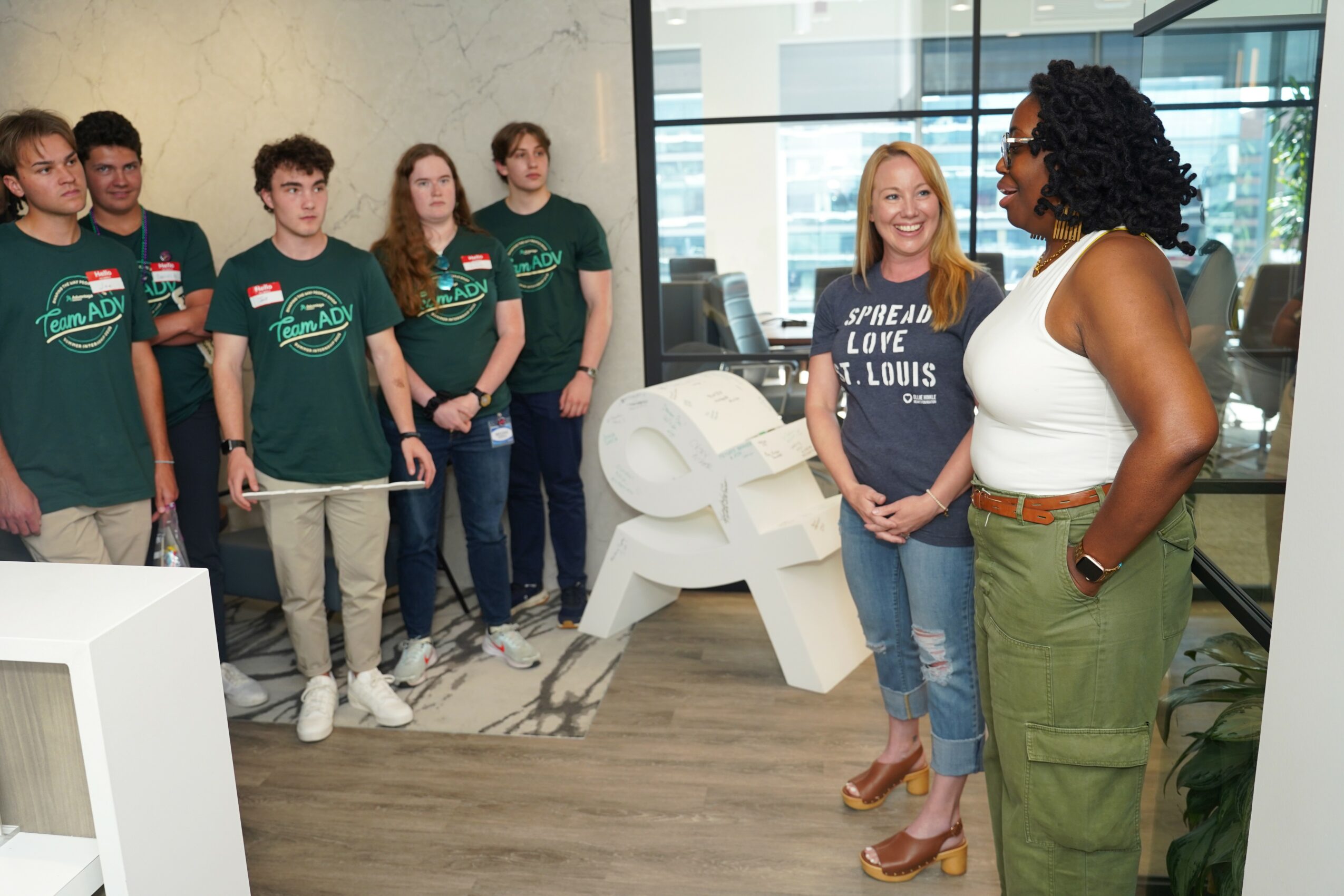 Group of young people listening to a woman speaking indoors, with a large white sculpture and glass windows behind them.
