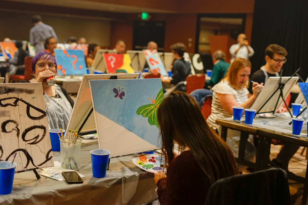 People participating in a painting event, with canvases, brushes, and blue cups on tables in a large room.