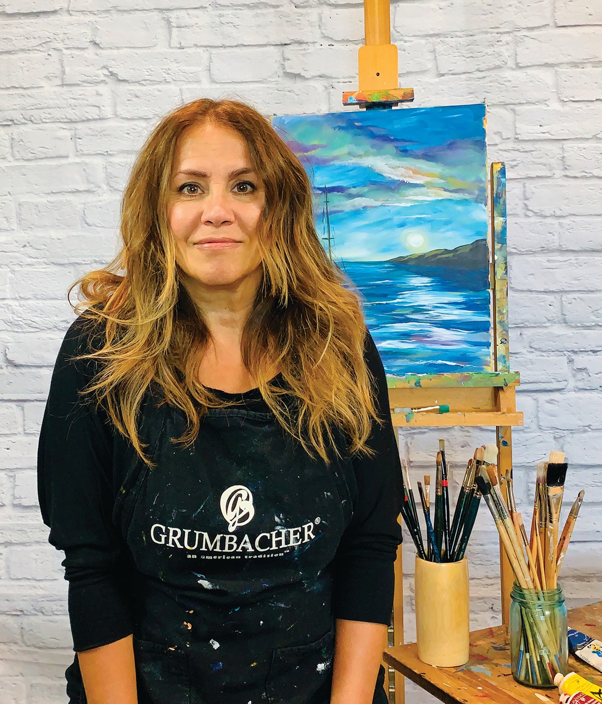 Woman with long wavy hair standing in front of an easel with a landscape painting, in a studio with a white brick wall.