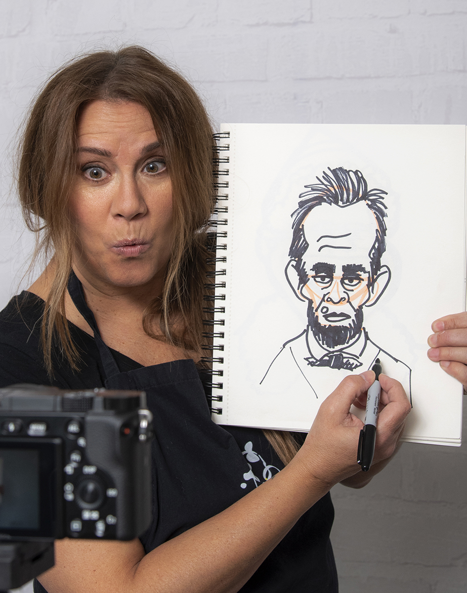 Woman with brown hair holding a sketchbook with a portrait drawing, camera in foreground, white brick wall background.