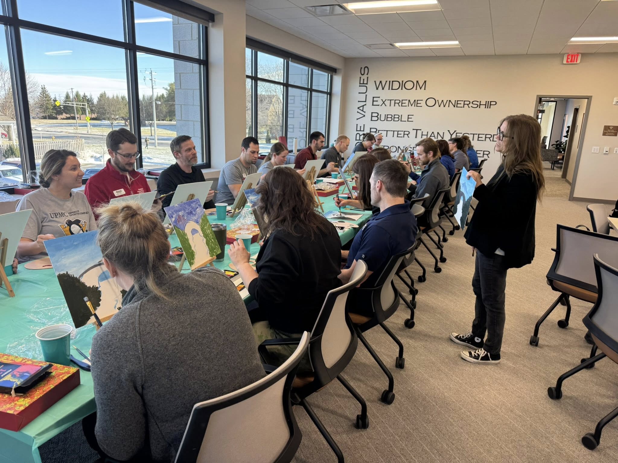 Group of people seated around a conference table in a room with large windows, listening to a woman standing and speaking.