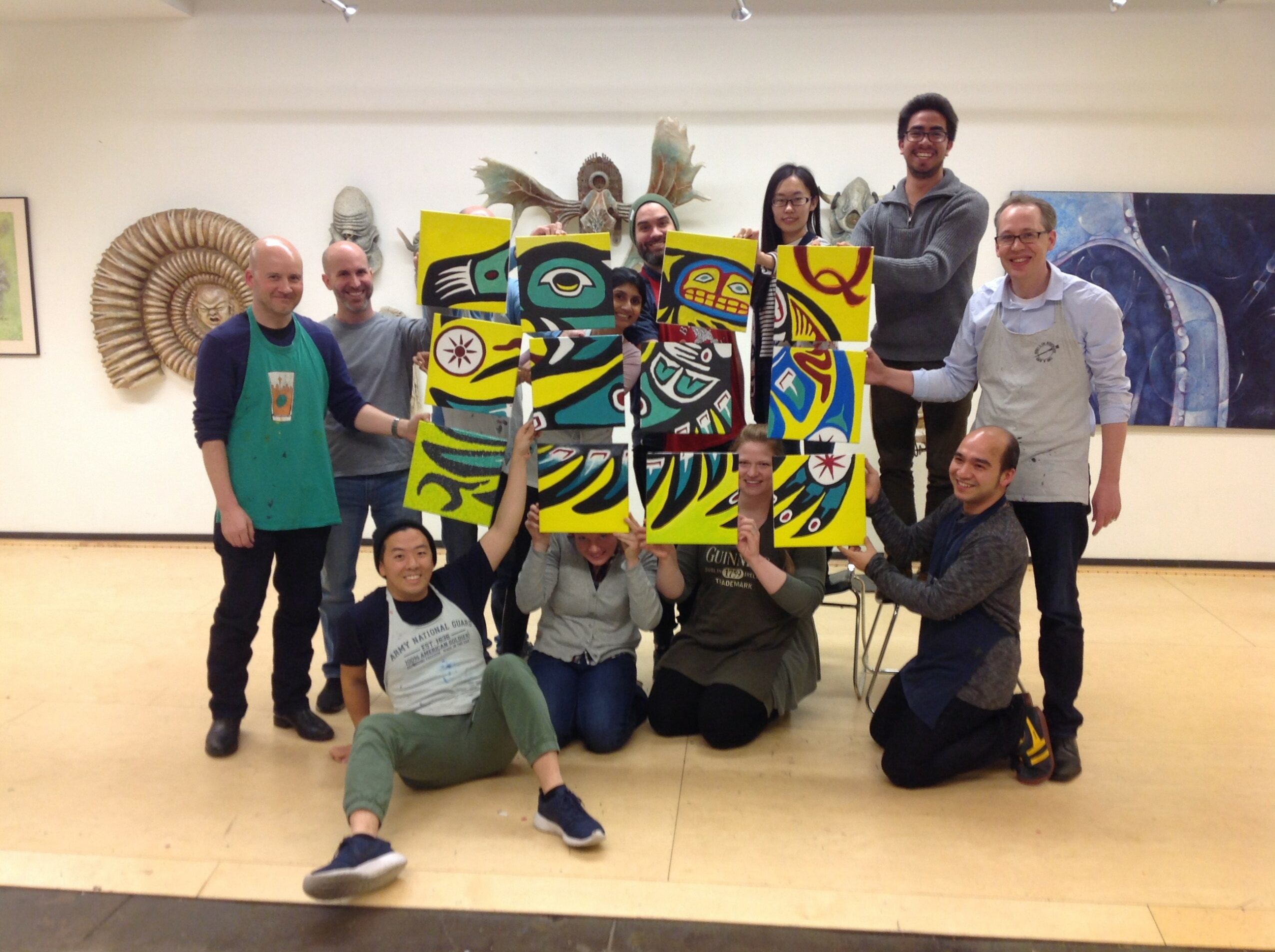 Group of ten people in an art gallery, holding colorful abstract paintings, some standing and some sitting, with artwork on walls behind them.