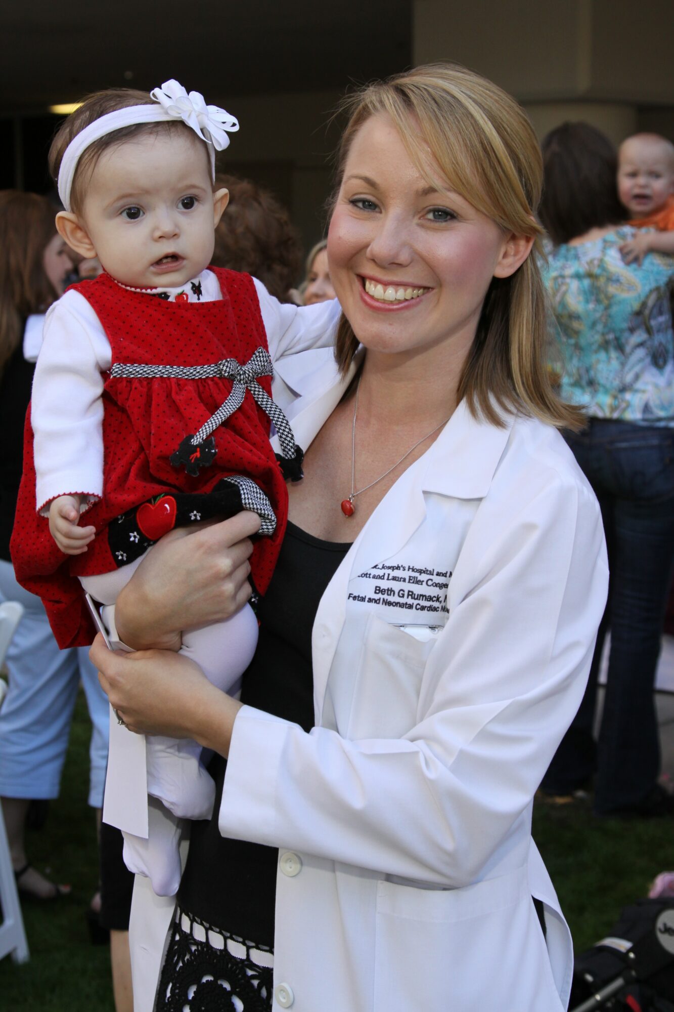 A woman in a white coat holding a young girl in a red dress with a white headband, smiling outdoors.