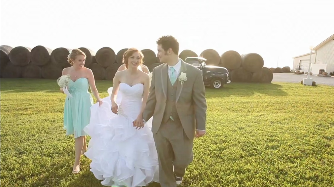 Three people walking on grass: a bride in a white dress, a groom in a suit, and a girl in a light blue dress.