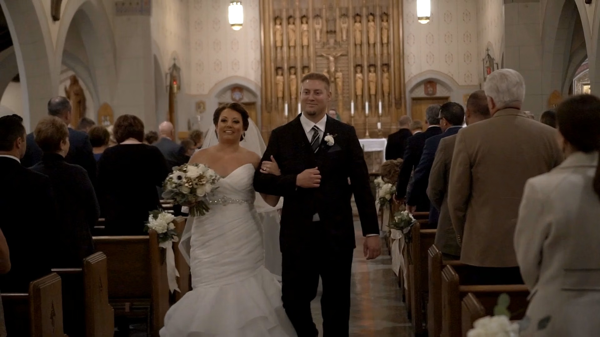 Bride and groom walking down the aisle in a church, surrounded by guests, bride holding a bouquet, church interior visible.