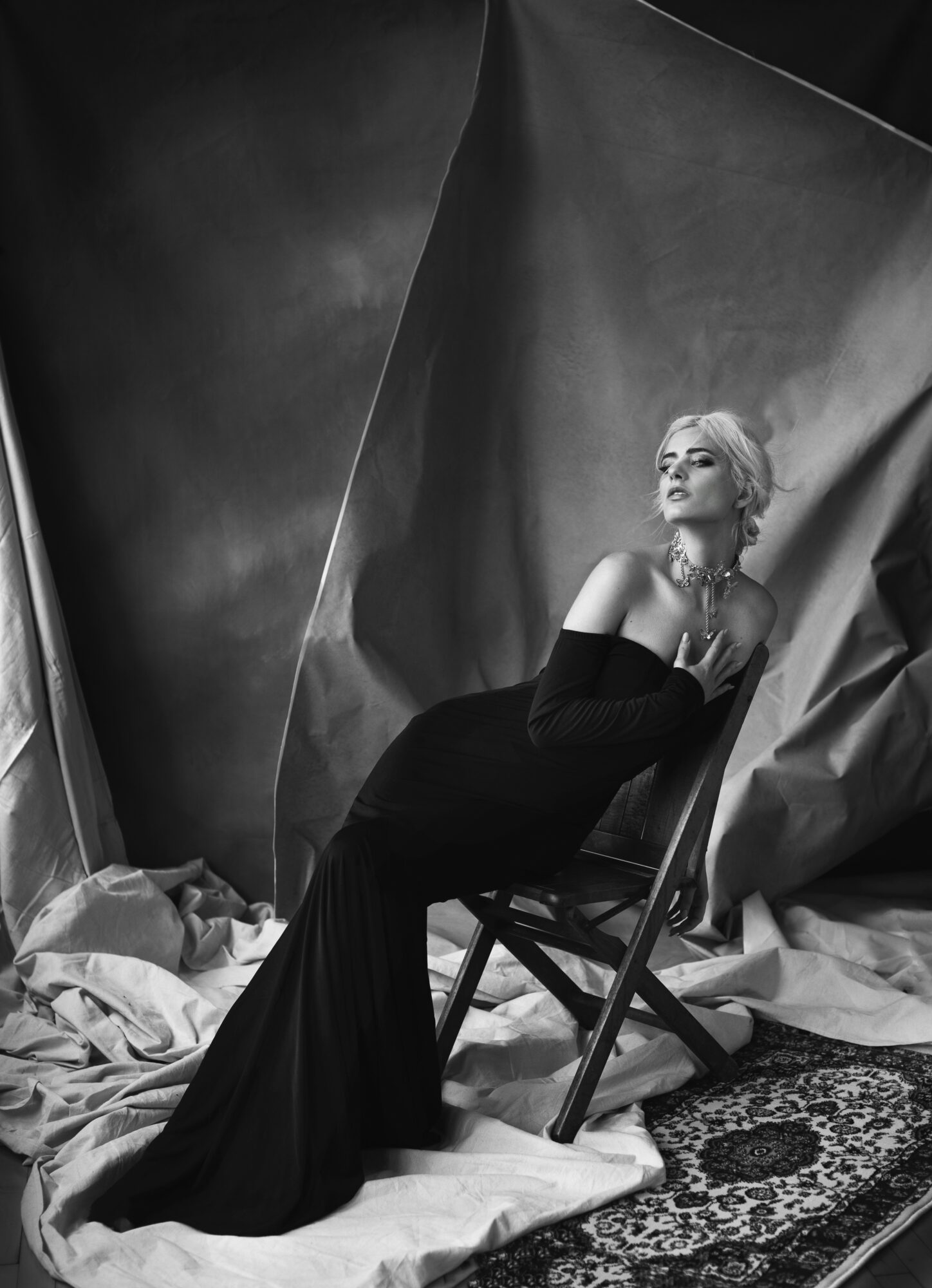 Woman in a long black dress sitting on a chair, surrounded by draped fabric, in a studio setting, black and white.
