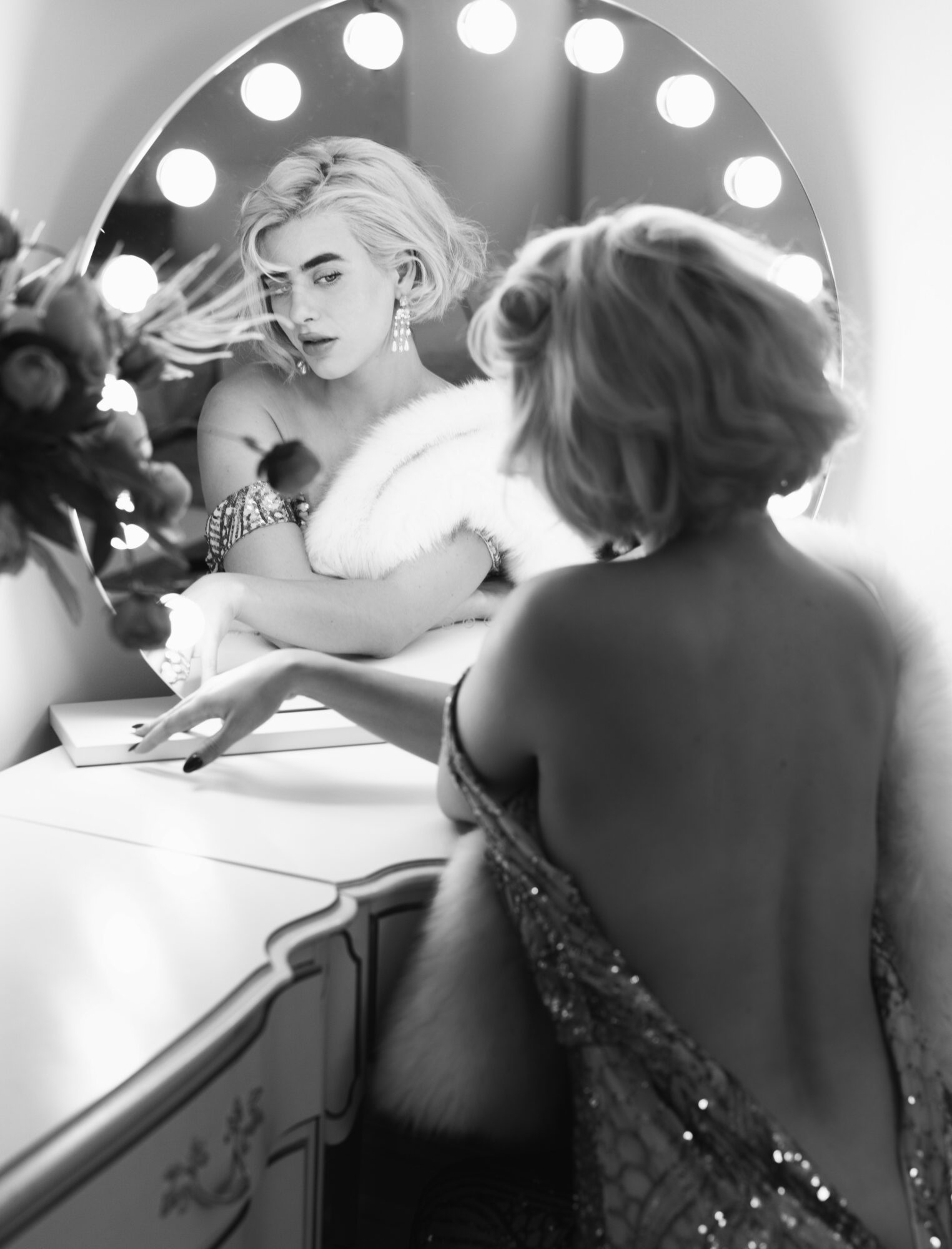 Two women in front of a mirror, one with short hair and the other with long hair, in a dressing room with lights.