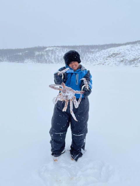 Child in winter clothing holding a snow-covered object in a snowy landscape with hills in the background.