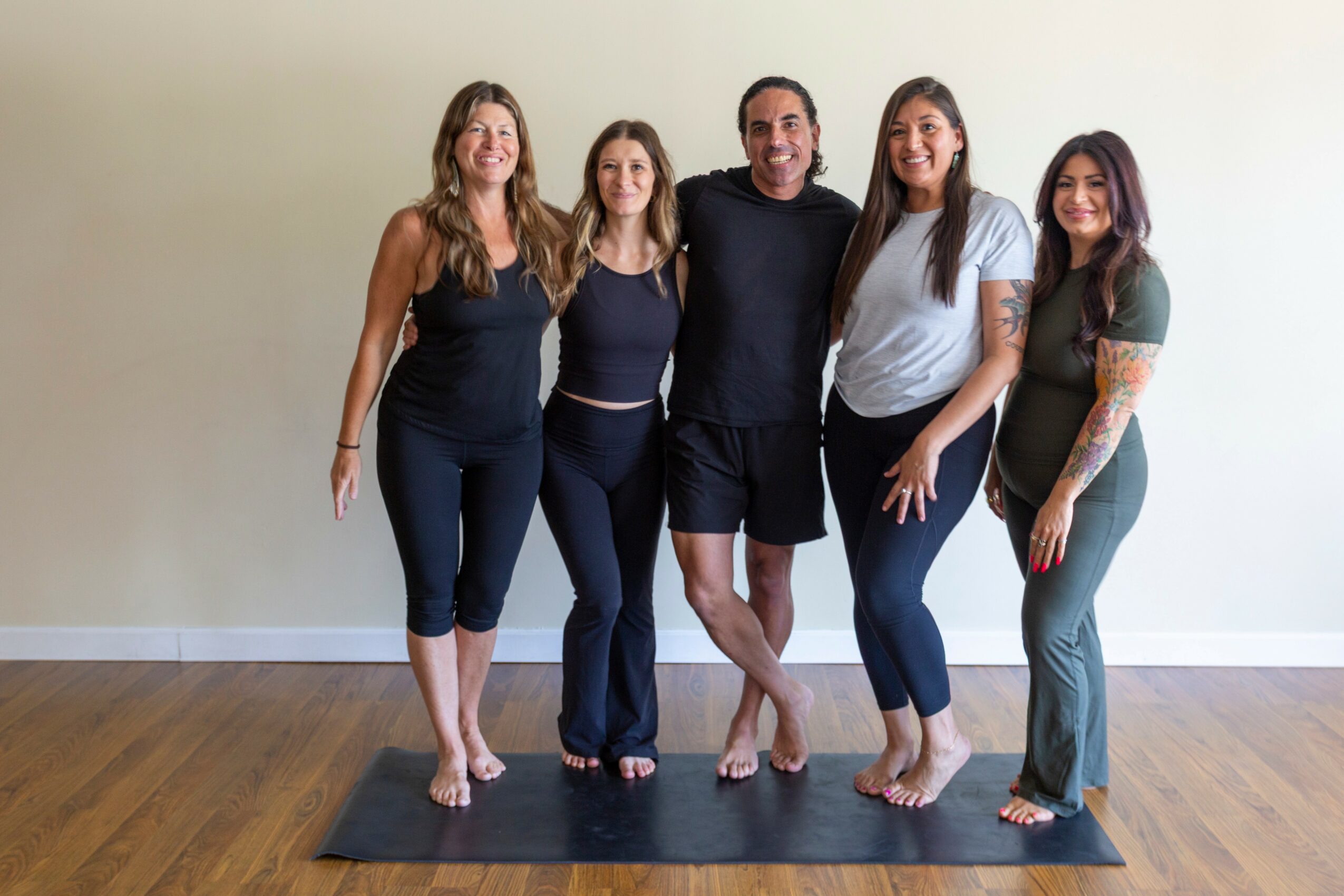 Five people standing together indoors, smiling, with a plain light-colored wall and wooden floor in the background.