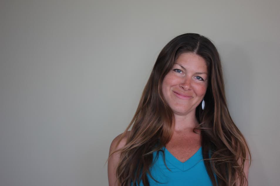 A woman with long brown hair smiling, wearing a blue top, standing against a plain light-colored wall.