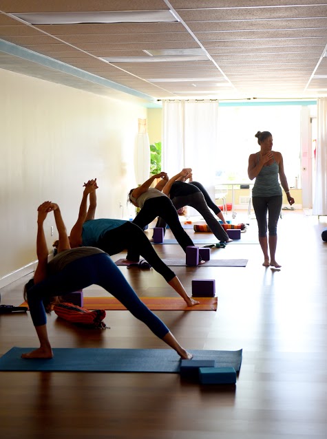 Group of people practicing yoga in a bright room with large windows, mats, and blocks, led by instructor. 20 words.