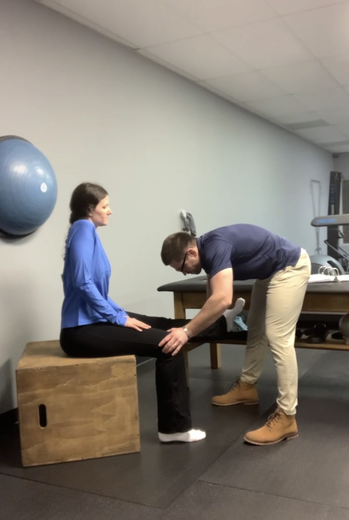 Person sitting on a wooden box with legs extended, another person leaning forward holding their ankles, in a room with a blue exercise ball on the wall.
