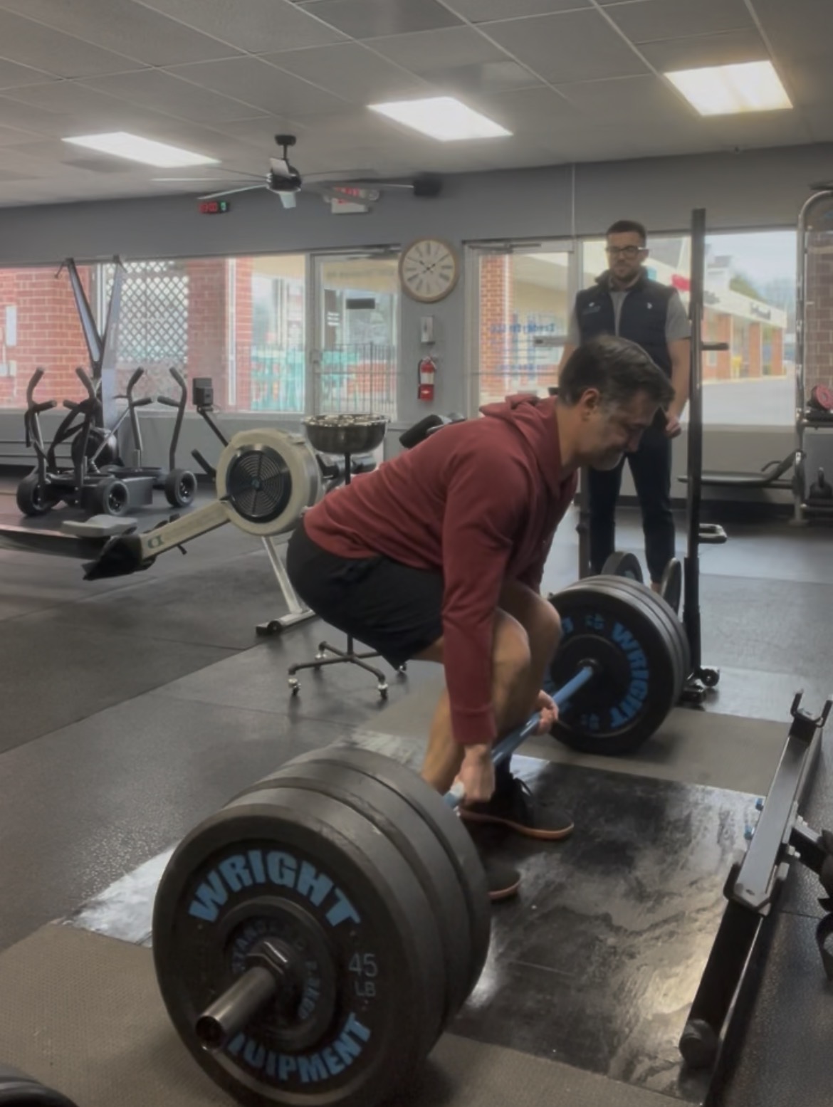 Person lifting a barbell with weights in a gym, with two people observing in the background, large windows, and gym equipment.