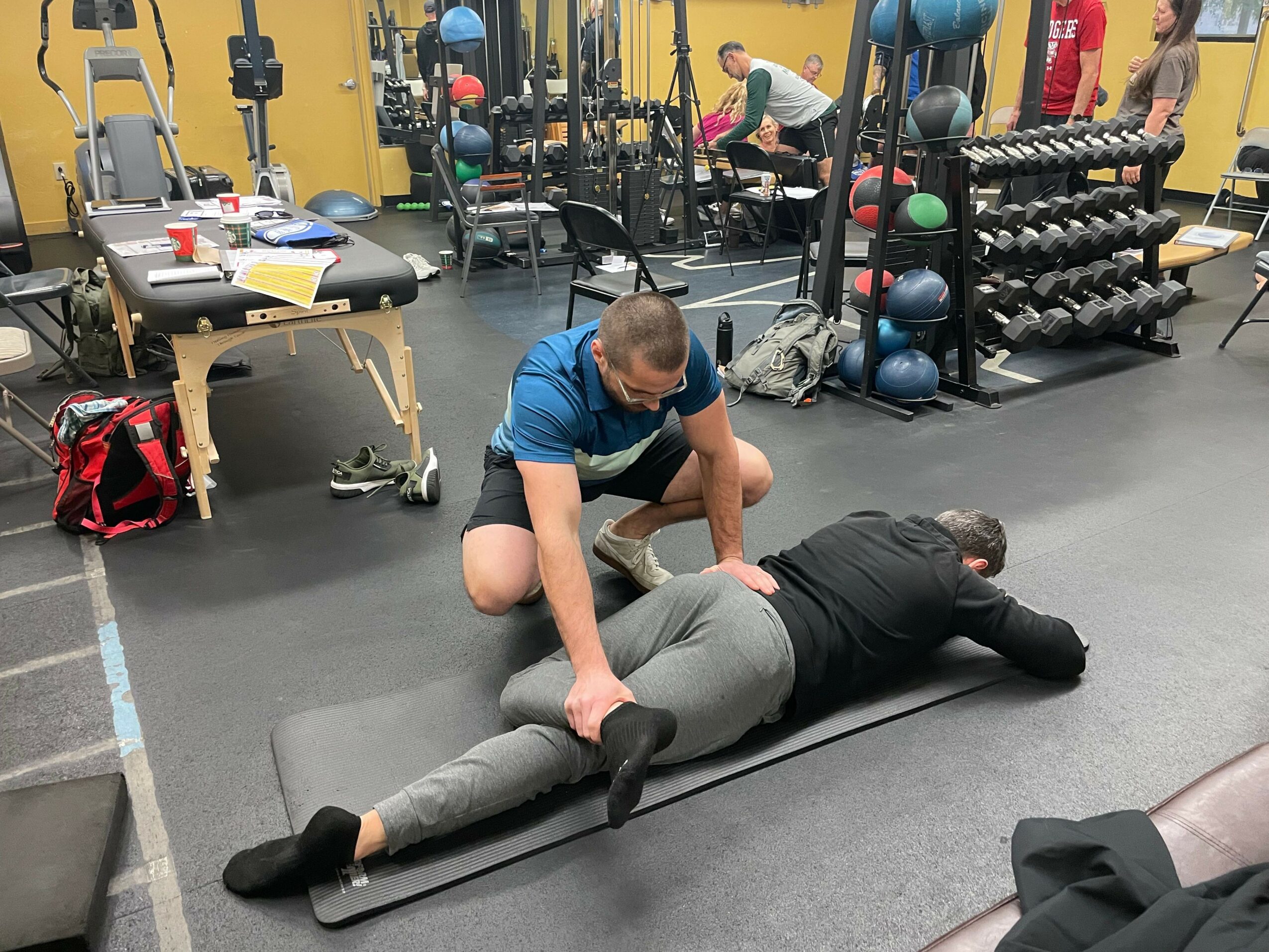 Person lying on a black mat, reaching for a barbell, in a gym with weight racks and equipment around.