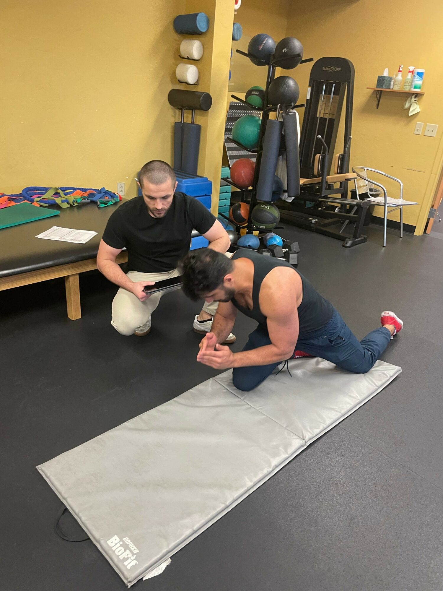 Two people practicing a fitness exercise on a padded mat in a gym, with gym equipment in the background.