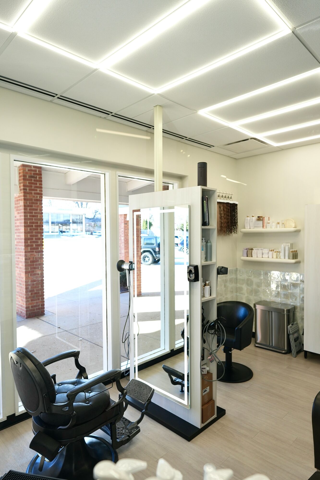 Salon interior with chairs, shelves, and a glass door, illuminated by ceiling lights, with a view outside through the door.