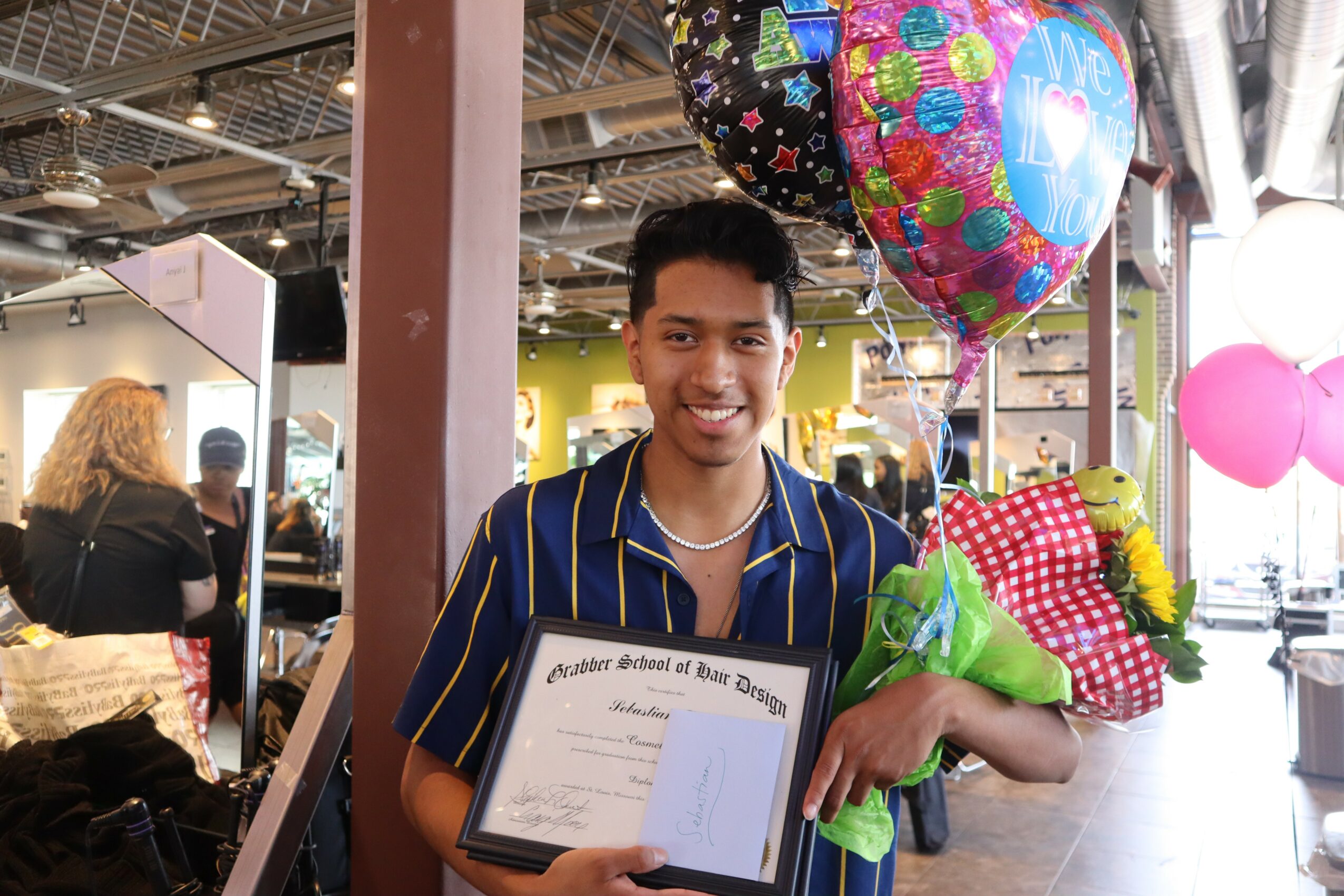 Young man holding a diploma and bouquet, smiling, in a decorated indoor space with balloons and people in the background.