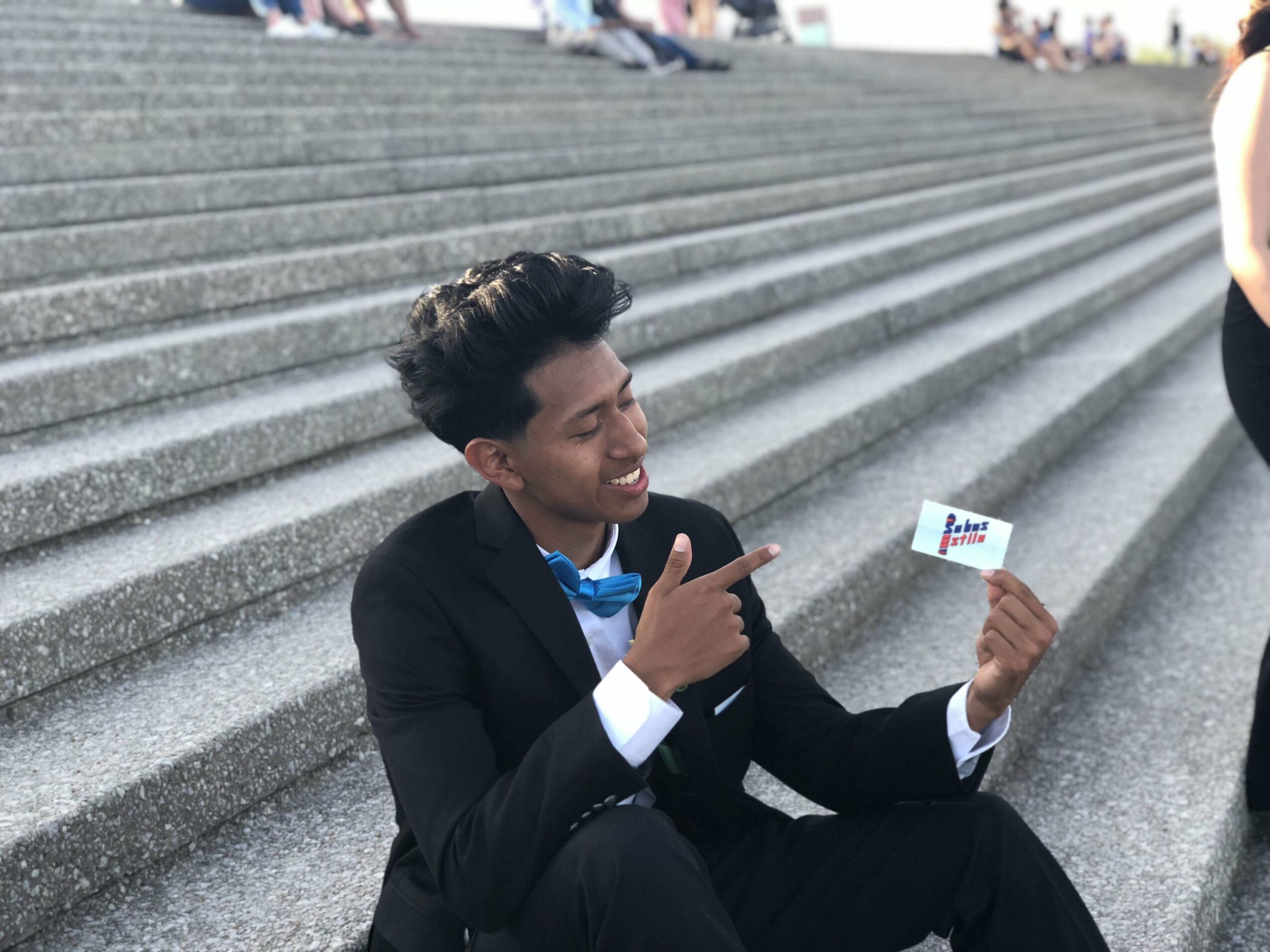 Young man in a suit sitting on stairs, holding a small card, smiling, pointing at it, outdoor setting