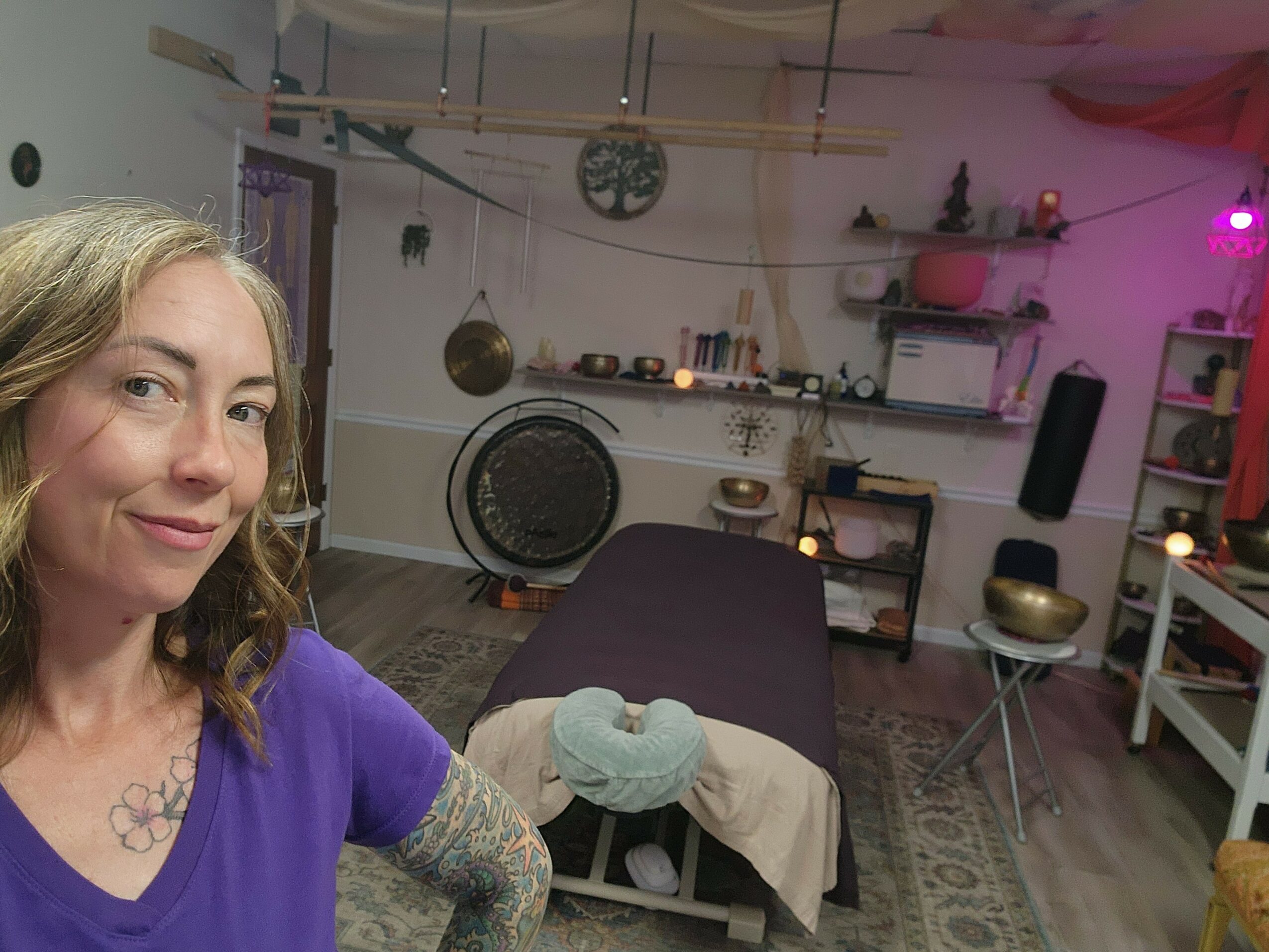 Woman with wavy hair in a purple shirt taking a selfie in a room with shelves, a massage table, and colorful lighting.