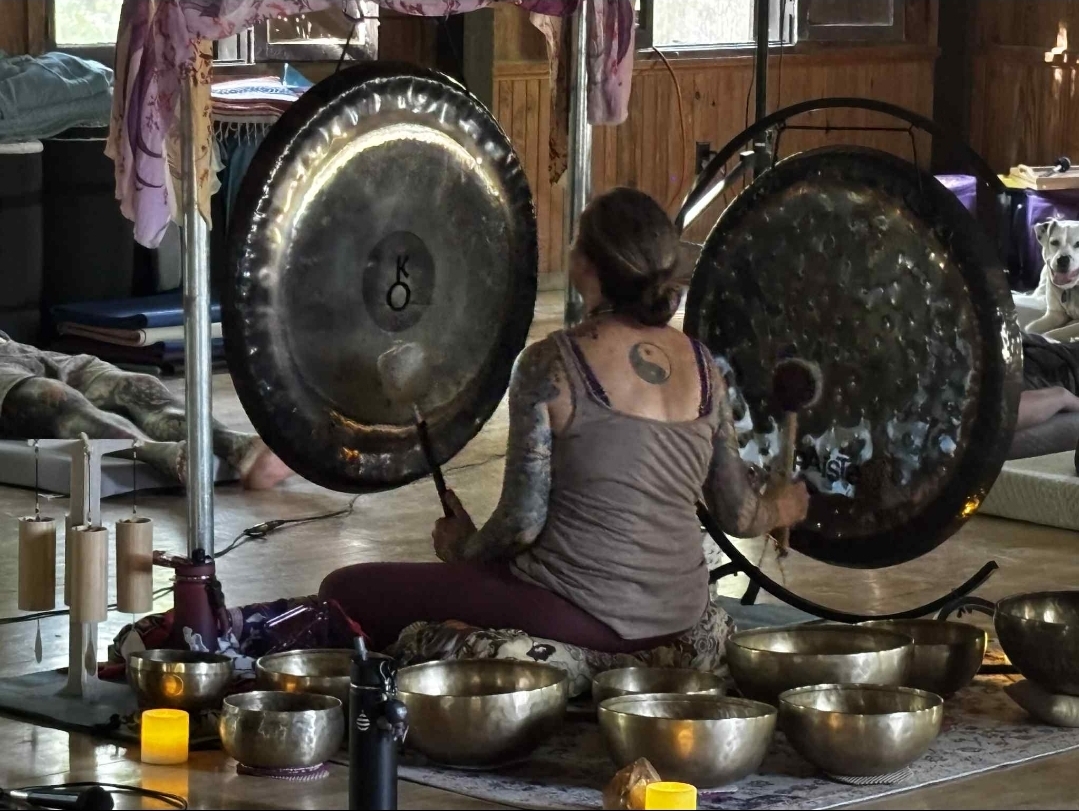 Person sitting cross-legged playing large gongs surrounded by smaller bowls and objects, in a wooden room.